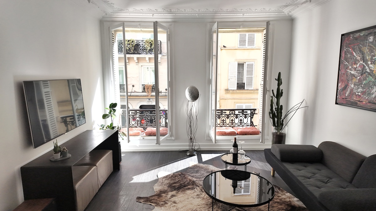 A bright living area is highlighted by large windows that open to a view of the street below. A dark sofa is paired with a round coffee table on a soft area rug. A wall-mounted television is positioned above a low, dark console table, while plants add a touch of greenery.