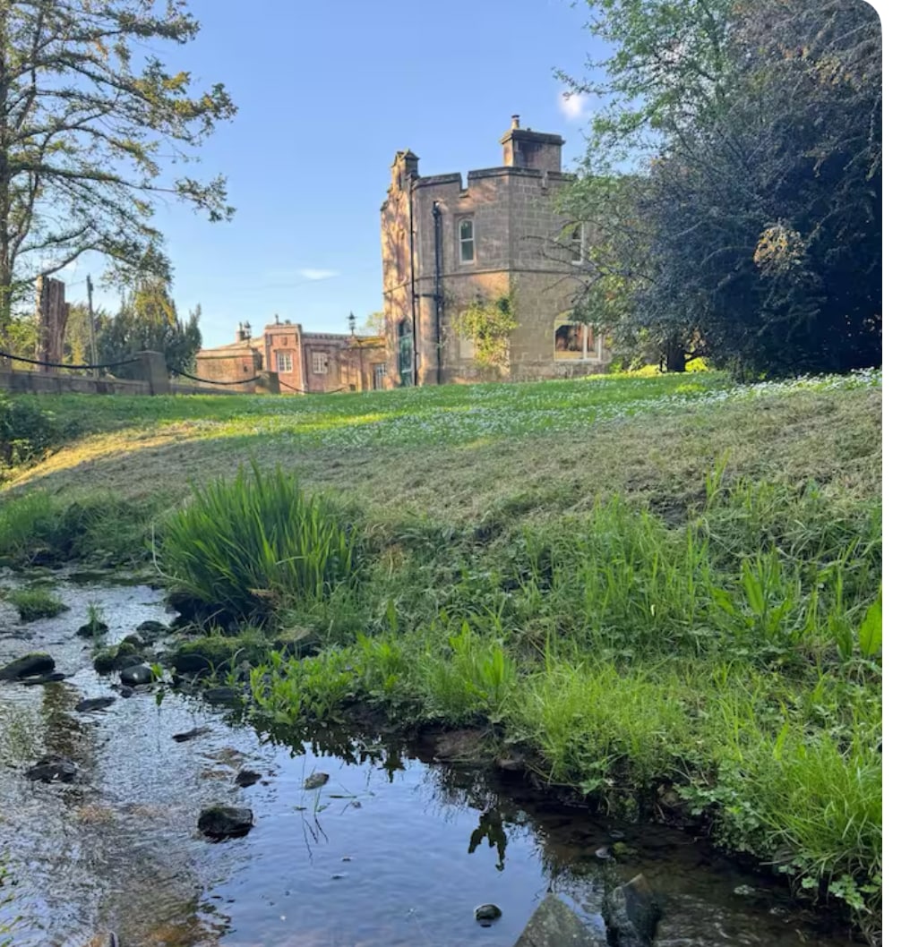 A serene view is presented featuring a babbling brook lined with lush greenery. Beyond the stream, a historic building can be seen, surrounded by well-maintained grounds under a clear blue sky. Wildflowers add a touch of color to the landscape.