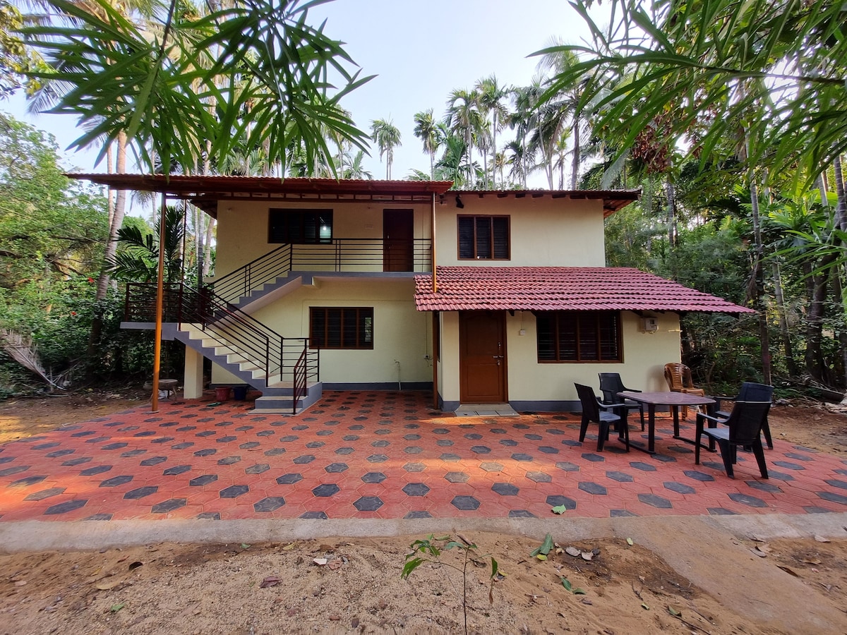 The property features a two-story structure with a red-tiled roof and a spacious outdoor area. A patio is visible in front, furnished with a table and chairs. Lush greenery surrounds the building, enhancing the natural setting of the property.