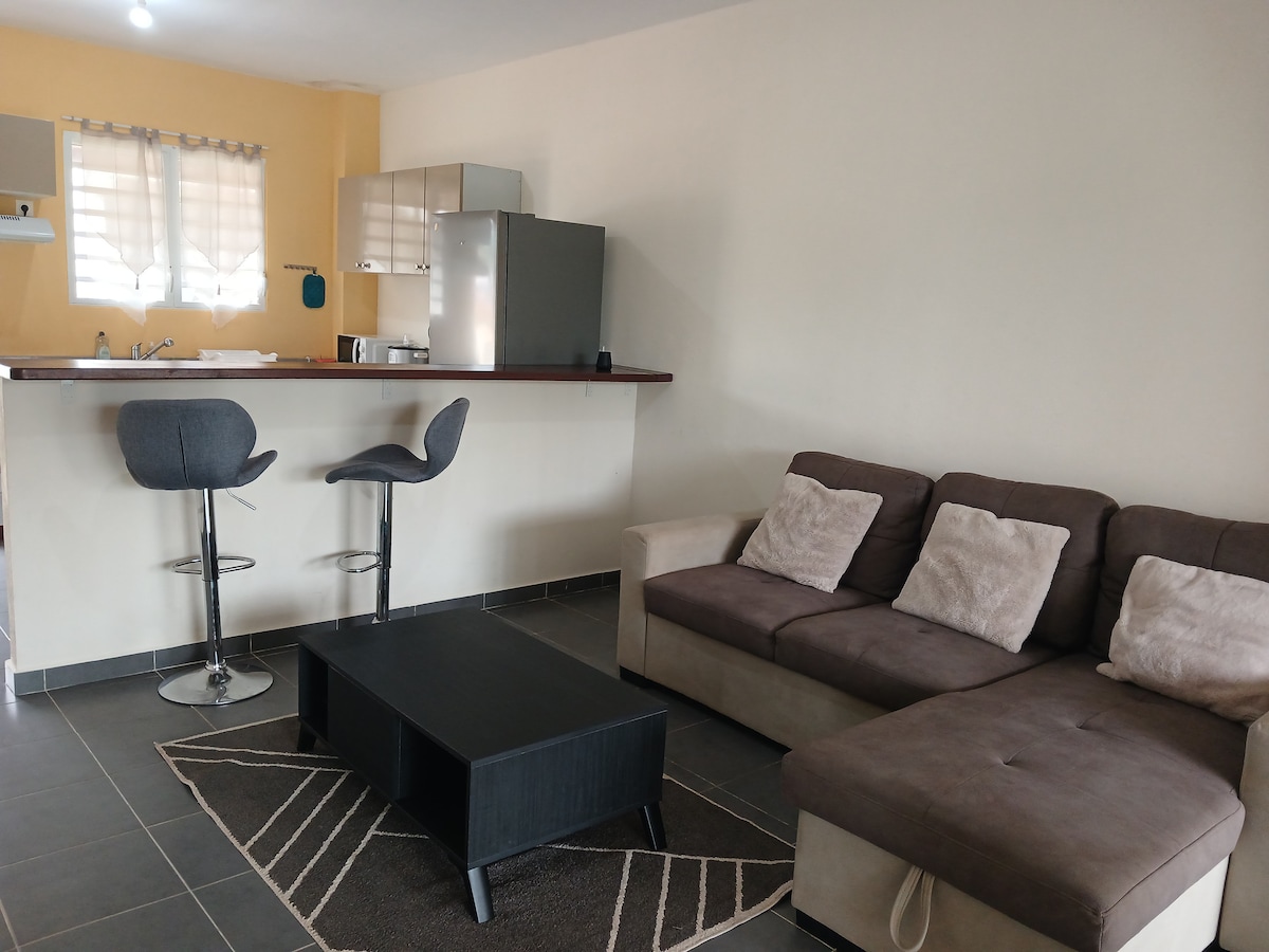 A comfortable living area features a sectional sofa with plush cushions and a low coffee table. Bar stools are positioned at a kitchen counter, which is visible in the background alongside modern appliances. Natural light enters through a window dressed with light fabric.
