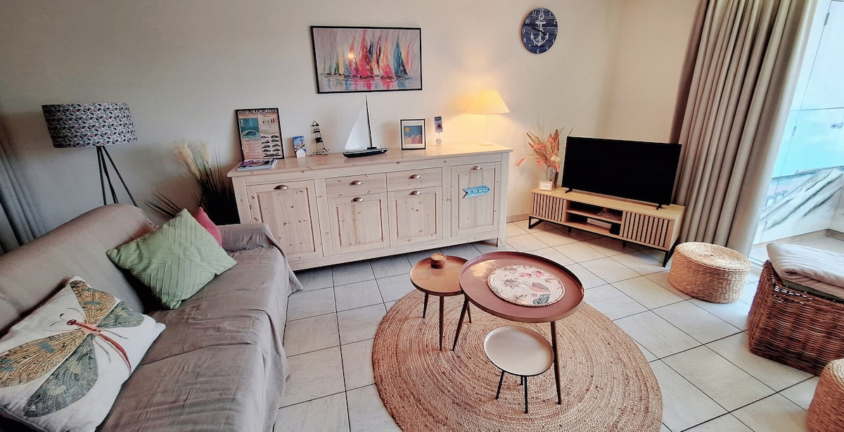 The living area features a light-colored sofa adorned with decorative pillows, accompanied by a round coffee table. A woven rug adds texture to the tiled floor. Modern entertainment is provided by a television on a low cabinet, with natural light filtering through floor-to-ceiling curtains.