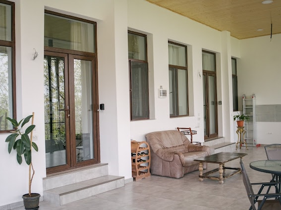 A spacious shared balcony features multiple large windows and a comfortable seating arrangement, including a leather sofa and wooden tables. Lush greenery is visible nearby, enhancing the outdoor connection of the space.