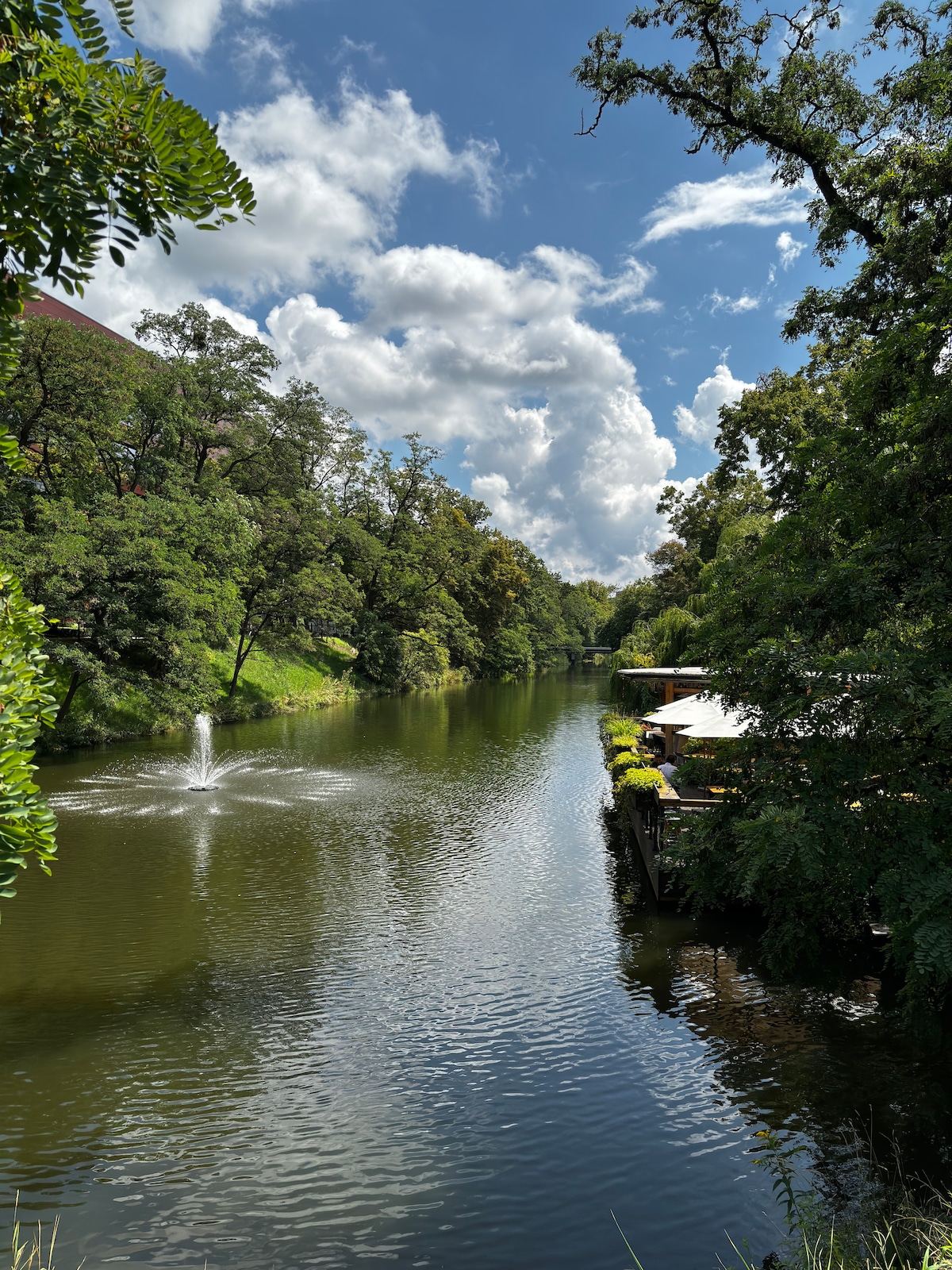 A serene view of a calm river is presented, with lush greenery lining the banks. A gentle fountain can be seen sending water into the air, while fluffy clouds drift across a bright blue sky. A nearby deck is partly visible, surrounded by trees.