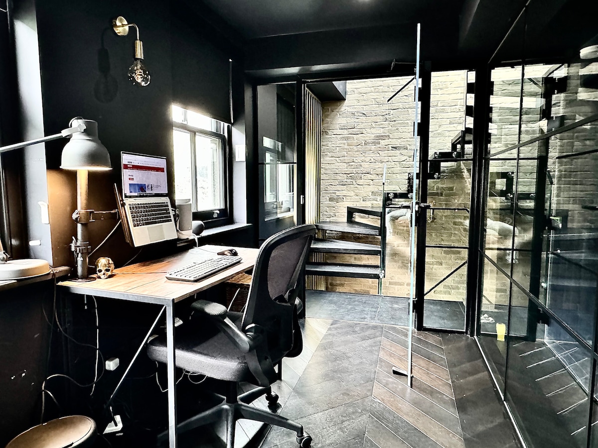 A dedicated office space features a modern desk with a computer and an ergonomic chair. Natural light enters through two windows, and acoustic glass partition walls provide a quiet atmosphere. A staircase is visible in the background, leading to other levels of the house.