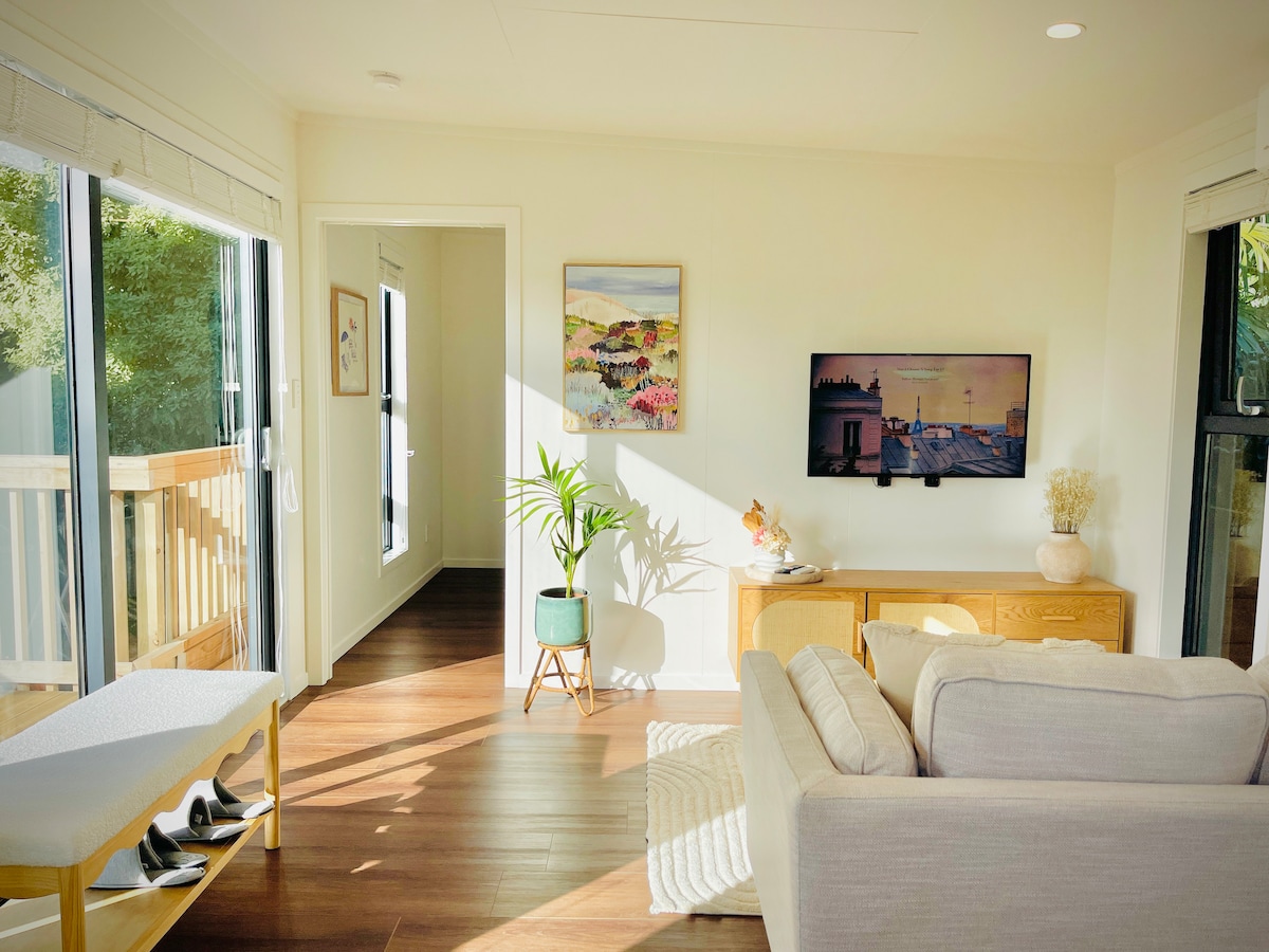 An open-plan living area is presented with natural light streaming in through large windows. A cozy couch faces a mounted television, while a light wood console holds decorative items. A small plant adds a touch of greenery, complementing the neutral color palette.