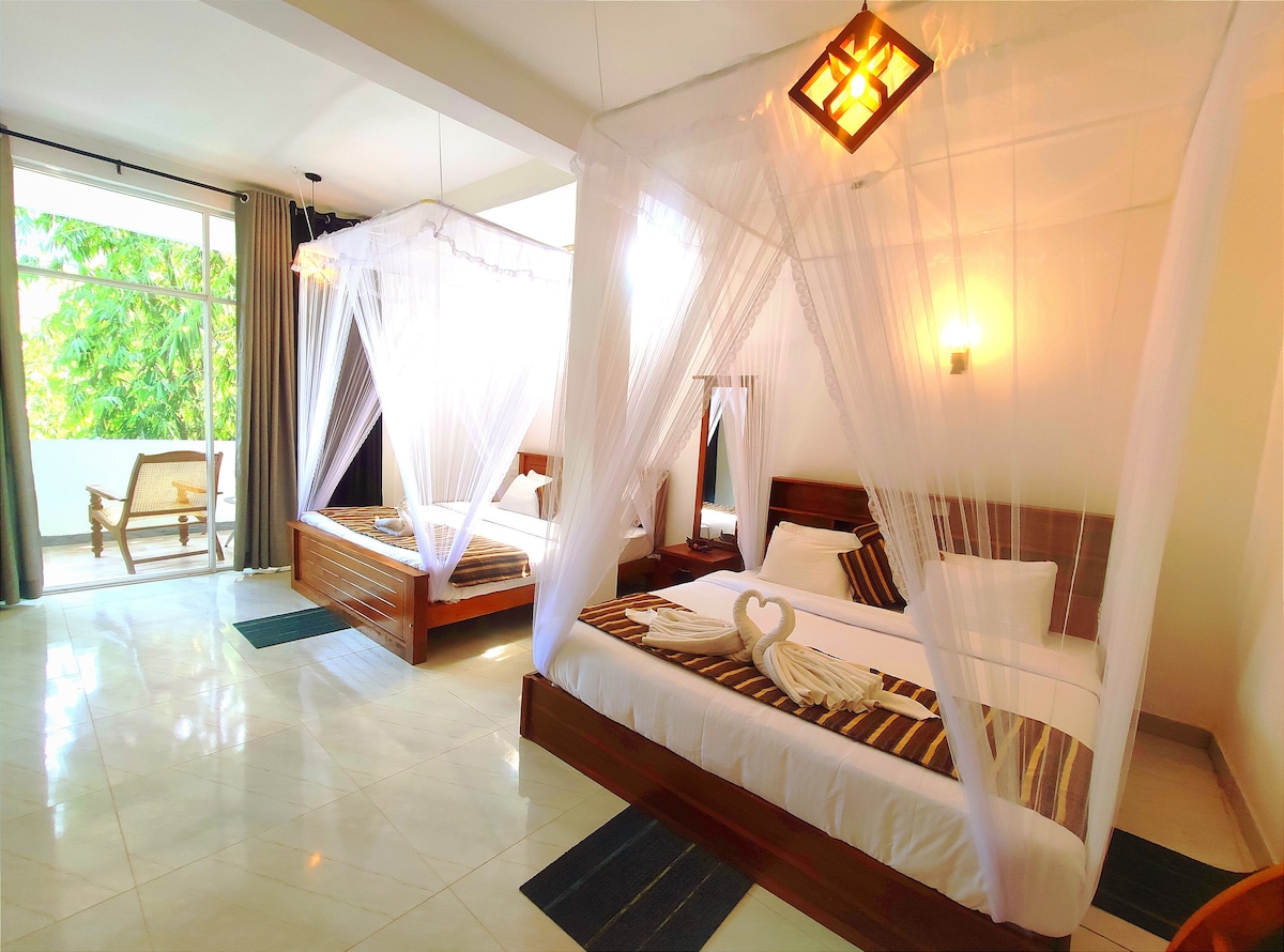 A spacious family room is furnished with two large beds, each draped with sheer mosquito netting. Natural light streams through large windows, providing views of lush greenery. Wooden furniture complements the serene ambiance, while decorative elements add a warm touch to the inviting space.