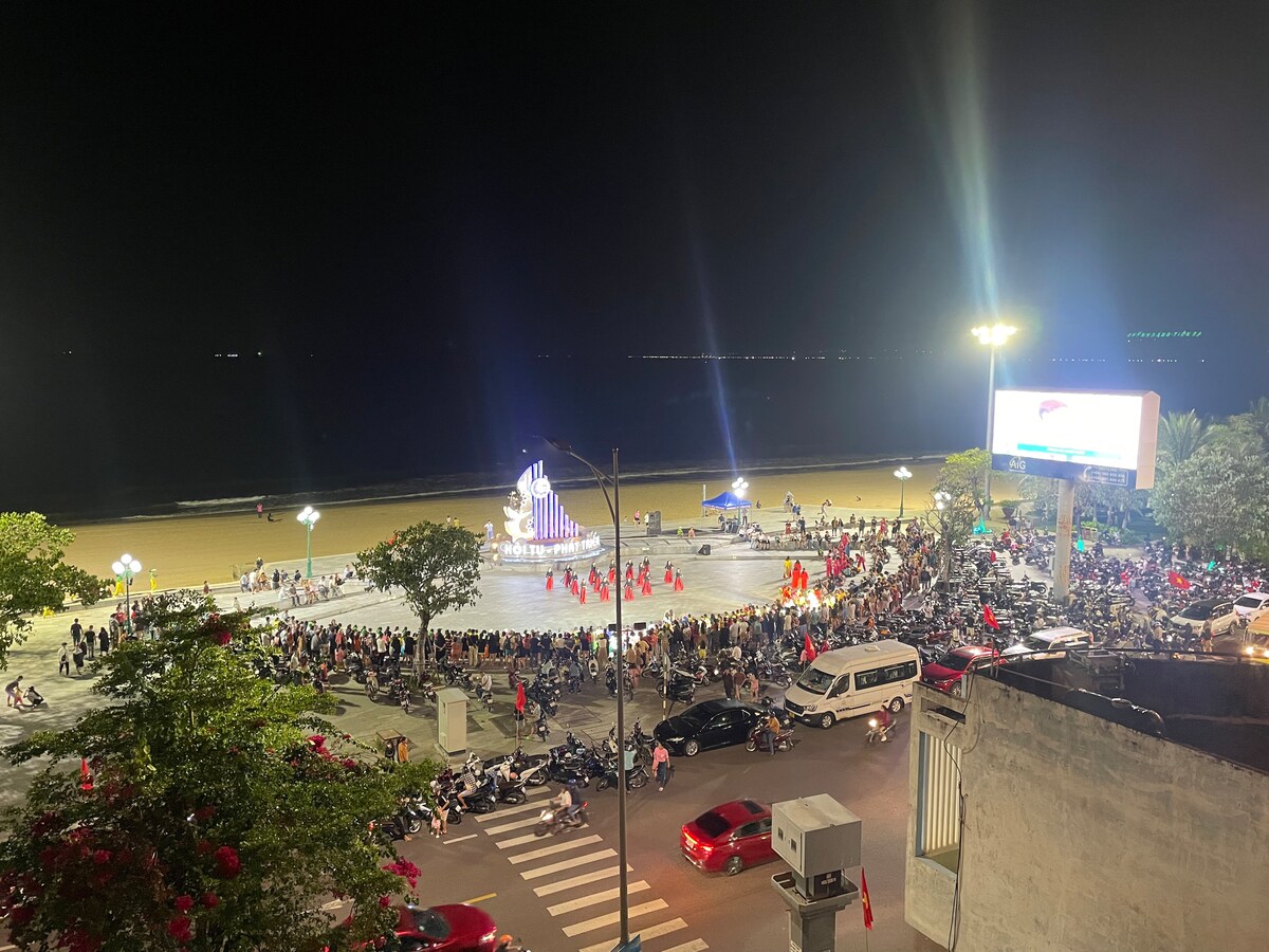 A lively outdoor gathering is depicted at night, with a performance area illuminated by bright lights. Spectators are seated and standing, while colorful performers in traditional attire entertain. The coastline is visible in the background, alongside parked vehicles and streetlamps lining the walkway.