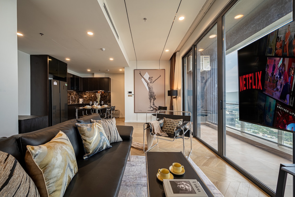 A contemporary living space features a comfortable sofa with decorative cushions, paired with a sleek coffee table. Large glass doors open to a balcony, allowing natural light to fill the room. A mounted TV displays streaming options, while a dining area is visible in the background.