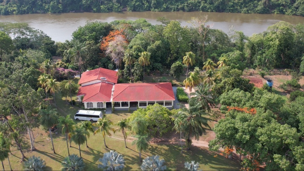 Aerial view of a spacious house nestled among lush greenery, showcasing a red-tiled roof and large windows. A river is visible in the background, surrounded by trees. The landscape includes well-maintained gardens and a mix of tropical plants, offering a serene natural setting.