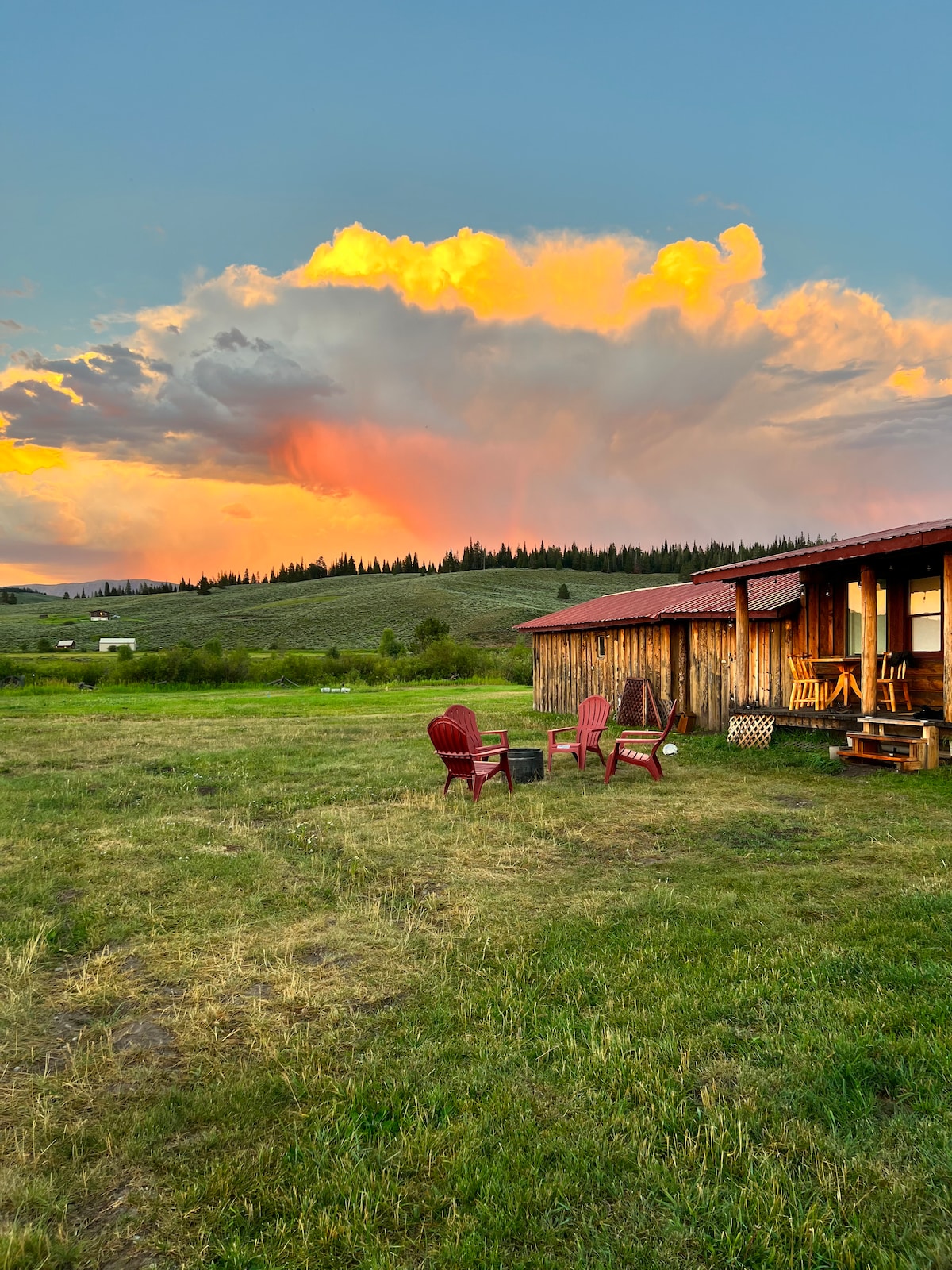 A scenic view of the studio cabin is highlighted by a colorful sunset in the background, with billowy clouds reflecting warm hues. In the foreground, four red outdoor chairs are arranged on a grassy area, inviting relaxation amidst the tranquil landscape of the surrounding mountains.