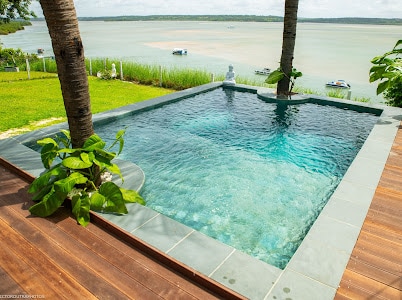A tranquil pool is set against a natural backdrop, with clear water reflecting the sky. Lush green plants and two palm trees frame the pool area, which is surrounded by wooden decking. A serene view of the lagoon is visible beyond the pool.