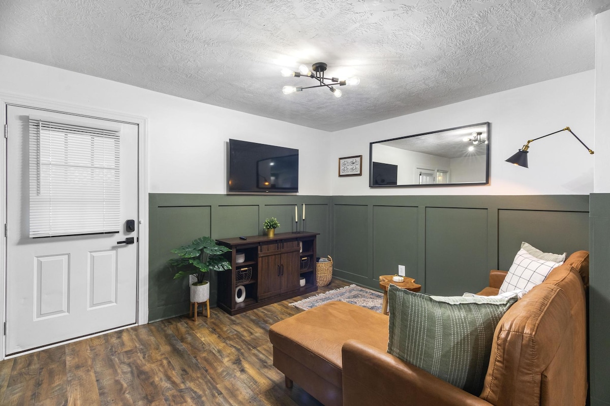 A comfortable living area features a brown leather sofa paired with patterned pillows. A flat-screen TV is mounted on the wall, with a decorative mirror reflecting light. The space is enhanced by greenery and a cozy area rug, alongside a door leading outside.