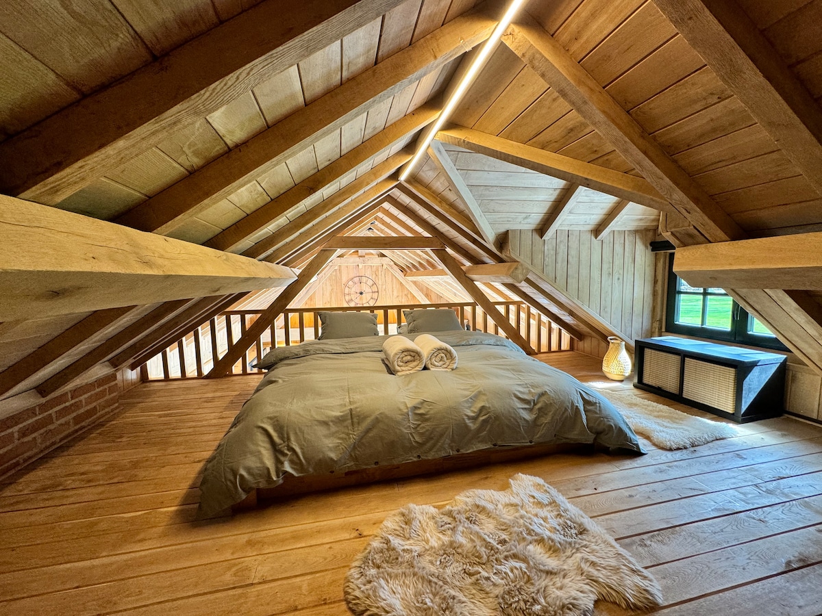 A charming mezzanine sleeping area is featured with a spacious bed dressed in soft linens. Wooden beams create an inviting ambiance while a warm light overhead adds to the cozy feel. A rug is placed on the wooden floor, enhancing comfort.