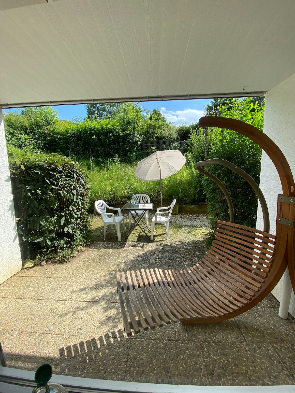 A welcoming outdoor space features a wooden swing seat and a small table with two chairs, shaded by an umbrella. Lush greenery surrounds the area, providing a serene atmosphere. Sunlight filters through, highlighting the textured surface of the stone patio.