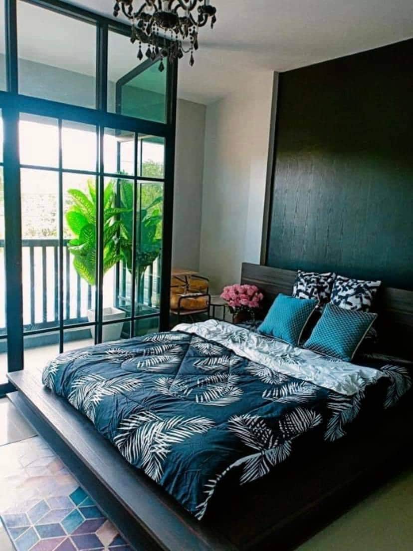 A modern bedroom features a spacious bed adorned with a patterned duvet and two decorative pillows. Large glass doors open to a balcony with vibrant greenery, while natural light enhances the room's neutral color scheme and highlights the stylish decor.