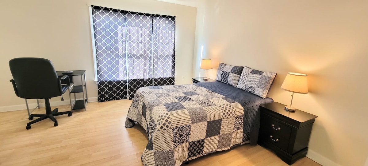 A master bedroom features a spacious bed adorned with a patterned quilt. Two bedside lamps provide soft lighting. A comfortable desk and chair are positioned against one wall, while large windows dressed with a decorative curtain allow natural light to fill the room.