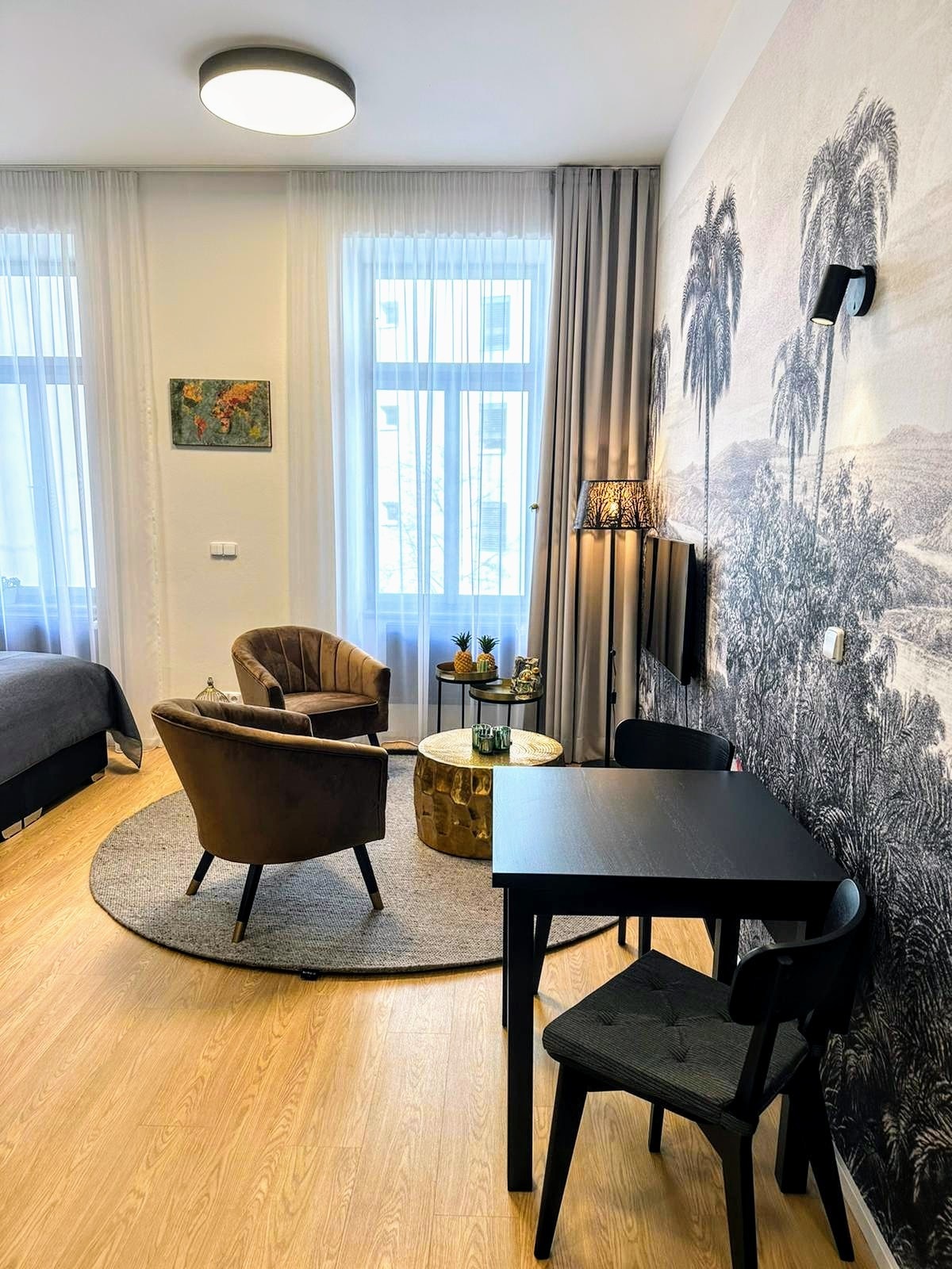 A cozy corner of the room features two upholstered chairs and a round table set on a circular rug. A decorative wall art piece and a lamp add character, while large windows allow natural light to enhance the neutral color palette.