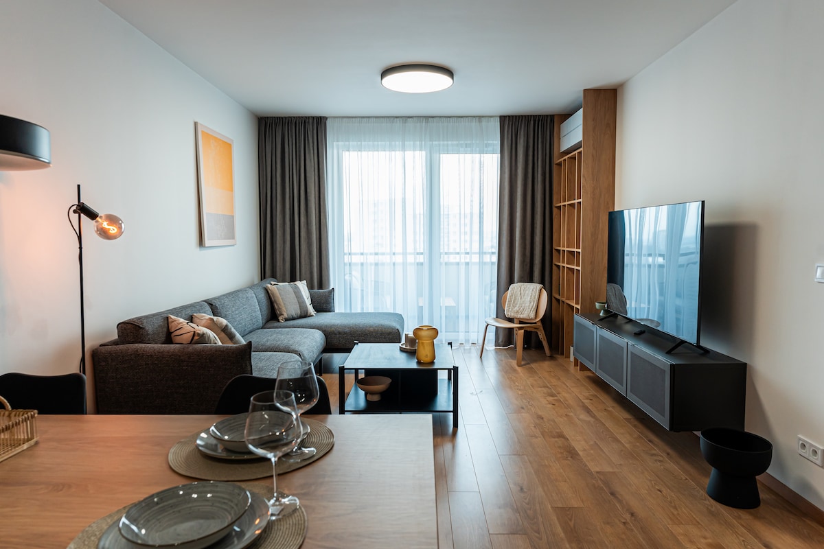 A modern living area features a large gray sectional sofa, a wooden coffee table, and a flat-screen TV mounted on a minimalistic unit. Natural light fills the space through a large window, complemented by sheer curtains. A dining table is set for four, enhancing the inviting environment.