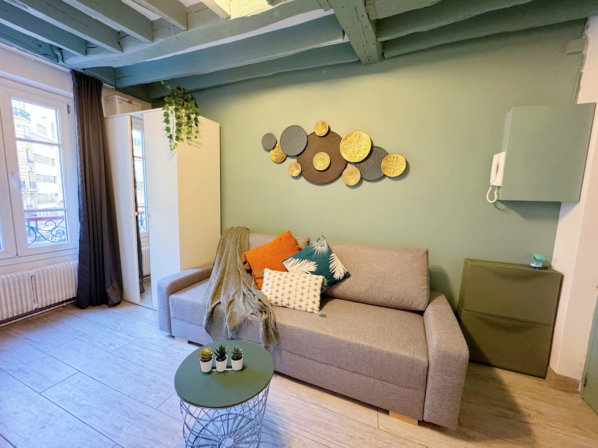 A comfortable lounge area is presented with a spacious gray sofa adorned with decorative cushions. A round green coffee table sits in front, complemented by small potted plants. The wall features a series of decorative plates, and natural light enters through a nearby window.