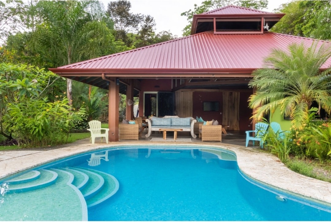 An outdoor seating area is situated beside a circular pool featuring a gentle staircase entry, surrounded by lush greenery and palm plants. The structure has a red roof and large open spaces that allow natural light to fill the interior.