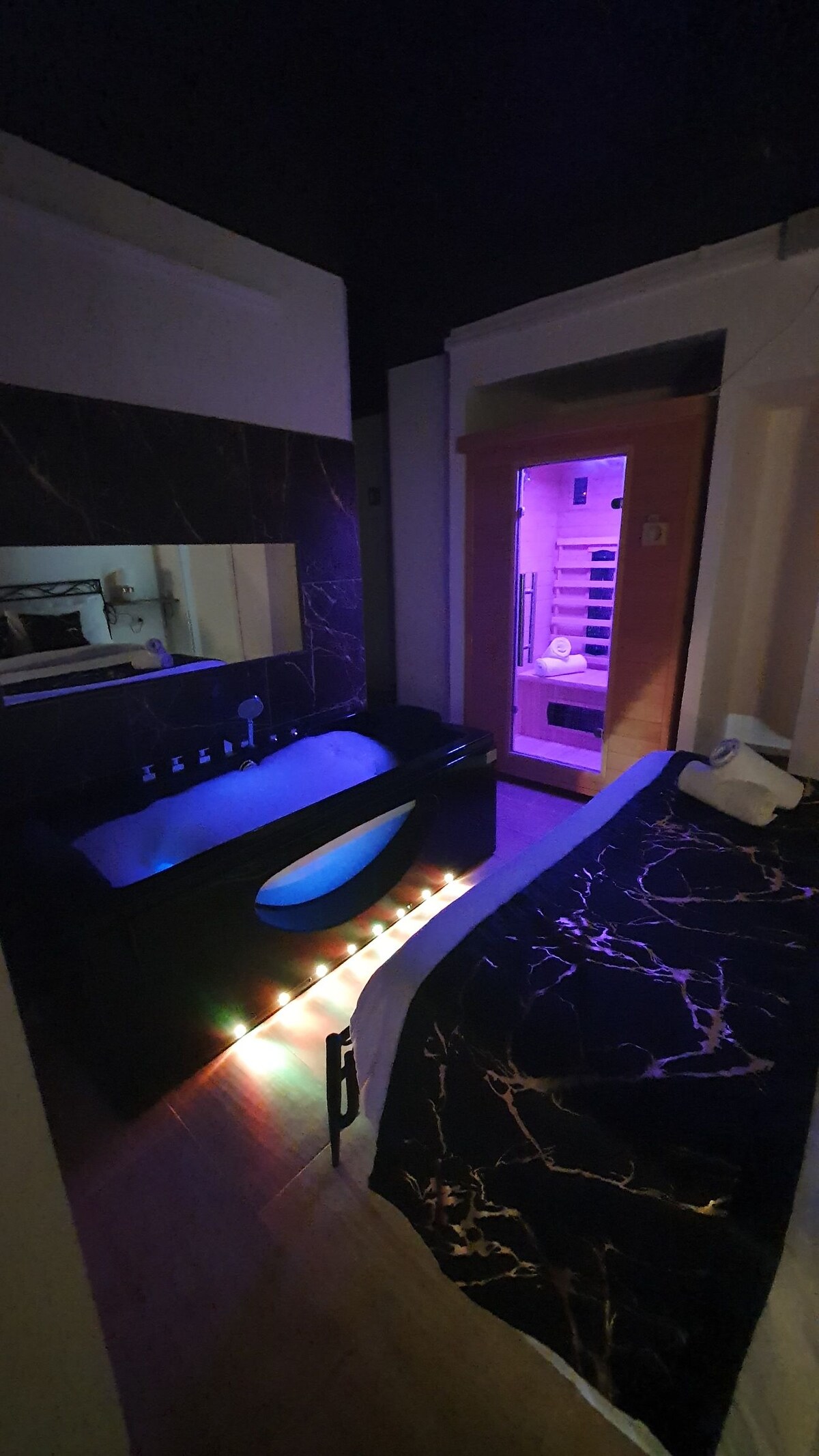 The image captures a modern space featuring a sleek black couch illuminated by soft lighting. A spacious bed with dark bedding is positioned beside a wooden sauna with softly glowing purple lights. A mirror reflects the contemporary design elements of the room.