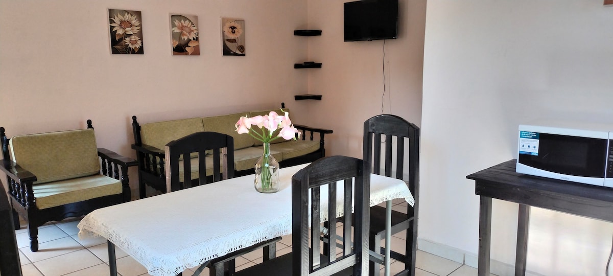 A spacious living area features a simple dining table surrounded by five chairs. A small black table sits nearby with a microwave. Two green sofas are positioned opposite the table. Wall-mounted shelves are visible, displaying framed artwork.