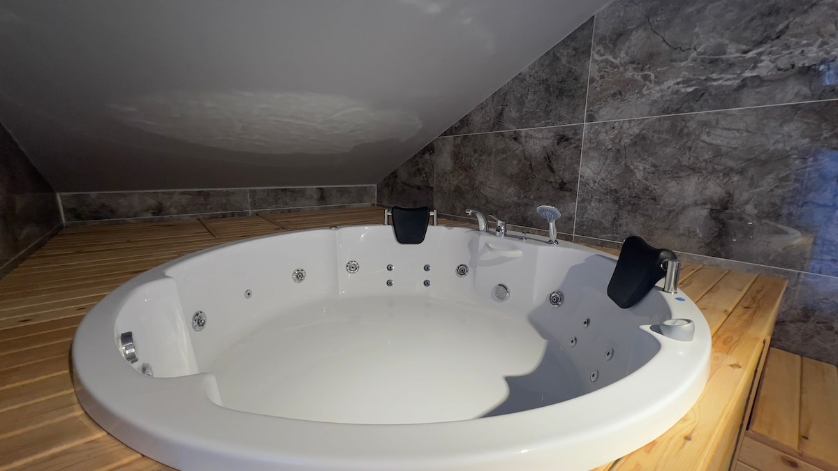 A spacious jacuzzi is presented in a softly lit corner with an angled ceiling. The tub features multiple jets and two black headrests, with wood paneling surrounding its base, creating a serene space for relaxation.