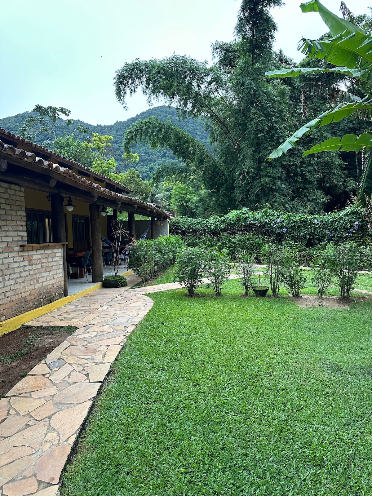 A well-maintained garden path winds through a green lawn, bordered by bushes and trees. The exterior of the accommodation features a brick wall, with a glimpse of an inviting outdoor seating area visible. The backdrop includes hills, enhancing the serene environment.