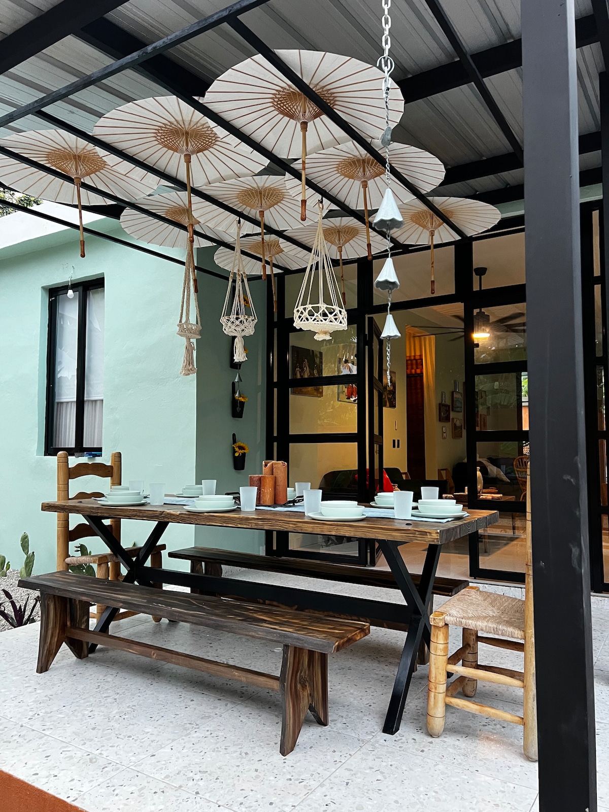 An outdoor dining area features a large wooden table set with dinnerware. Several hanging decorations, including round umbrellas and woven lanterns, create an inviting ambiance. The table is accompanied by a mix of wooden and bamboo chairs, with a light-colored wall and glass doors visible in the background.