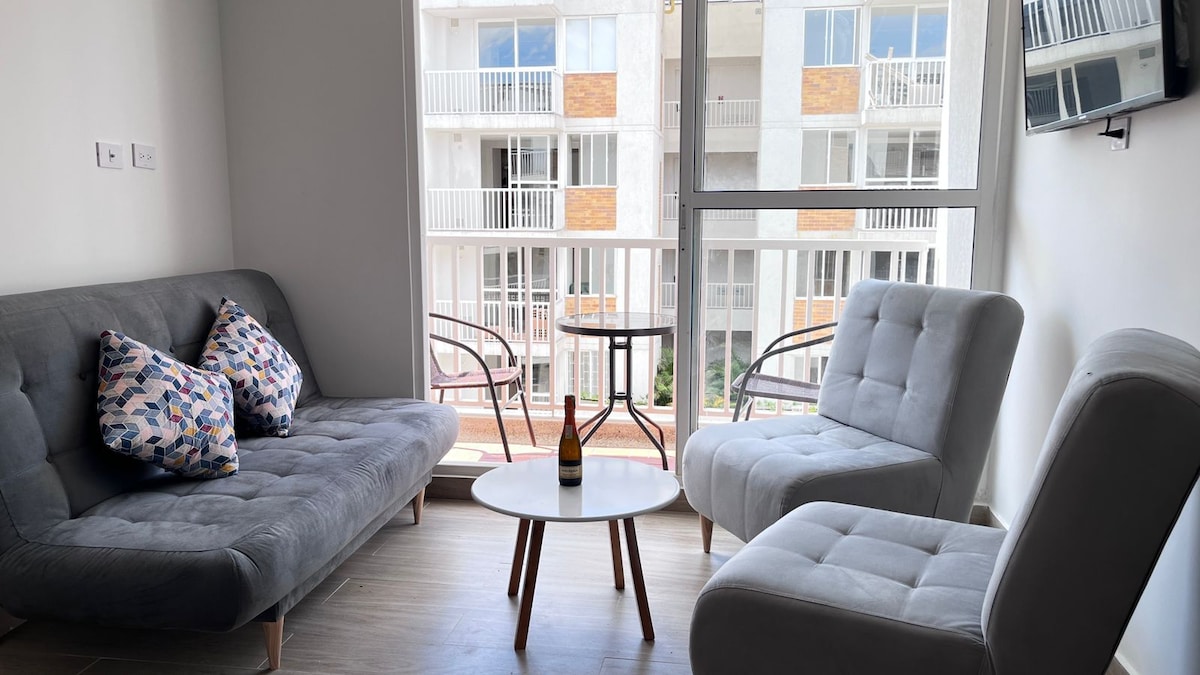 A modern living area is shown, featuring a pair of light grey armchairs and a sleek grey sofa adorned with patterned cushions. A small round white table is positioned in the center, while a large window offers views of the building facade outside.