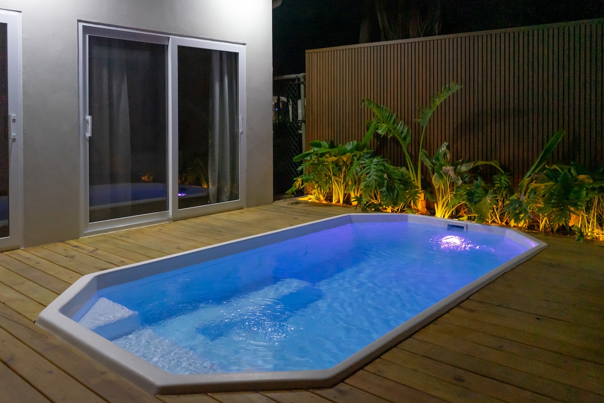 A serene pool area is showcased, featuring a rectangular pool with soft blue lighting and a wooden deck. Lush greenery is positioned around the pool, illuminated by subtle lights, creating a tranquil environment during the night.