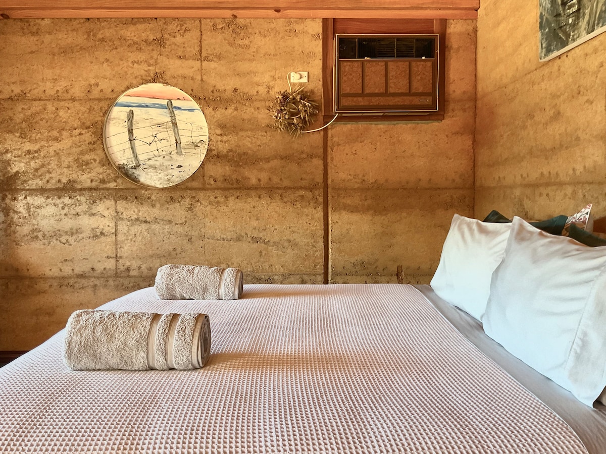 A spacious sleeping area is highlighted by a double-height timber ceiling and textured rammed earth walls. A comfortable bed is dressed in a light blanket, with neatly rolled towels placed at the foot. Artwork hangs above, and an air conditioning unit is mounted nearby.