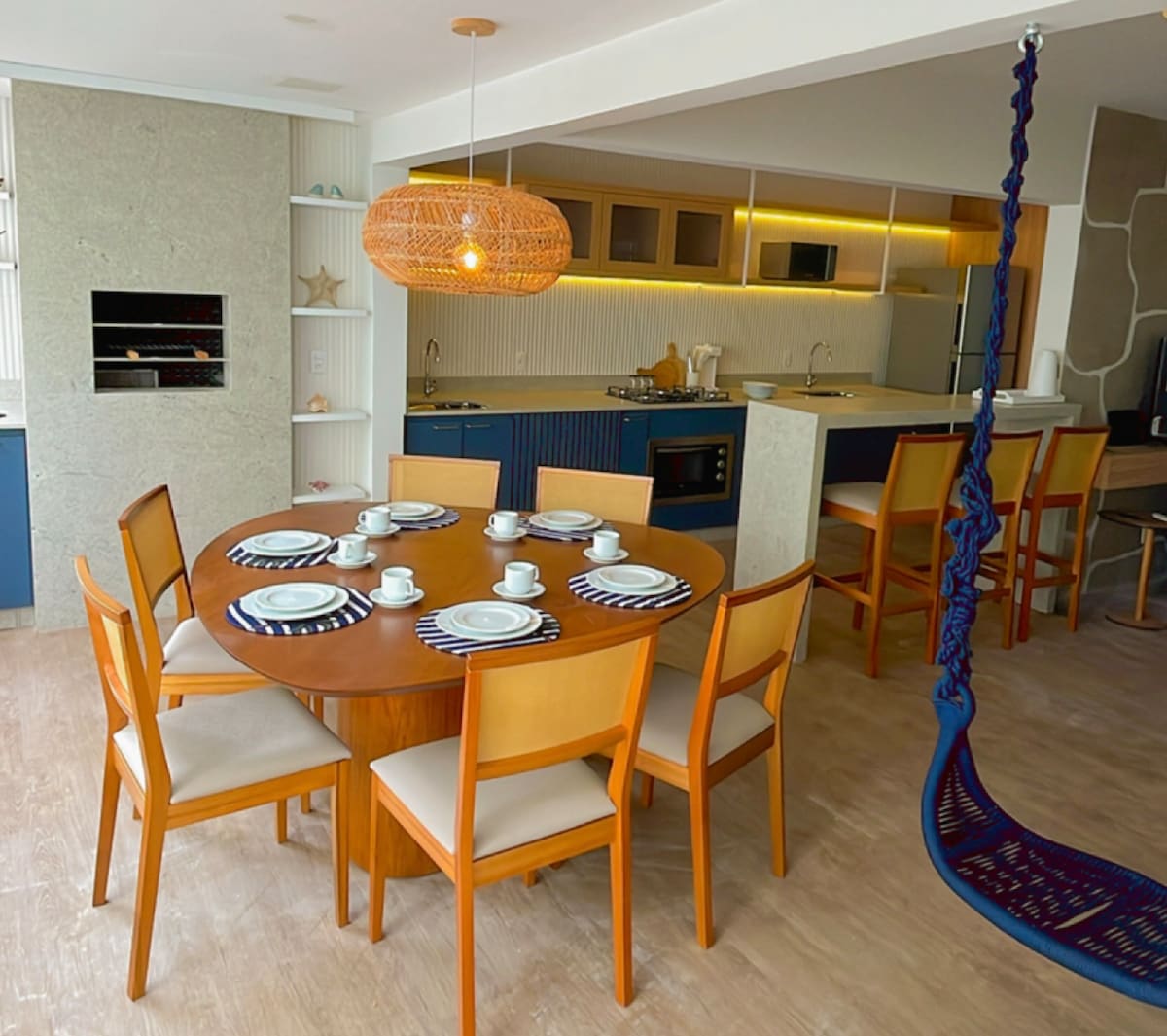 A dining area is presented with a round wooden table surrounded by six chairs. The table is set with plates and mugs. The modern kitchen is visible in the background, featuring sleek cabinetry and a built-in grill. A hammock occupies one corner, adding a relaxed touch.