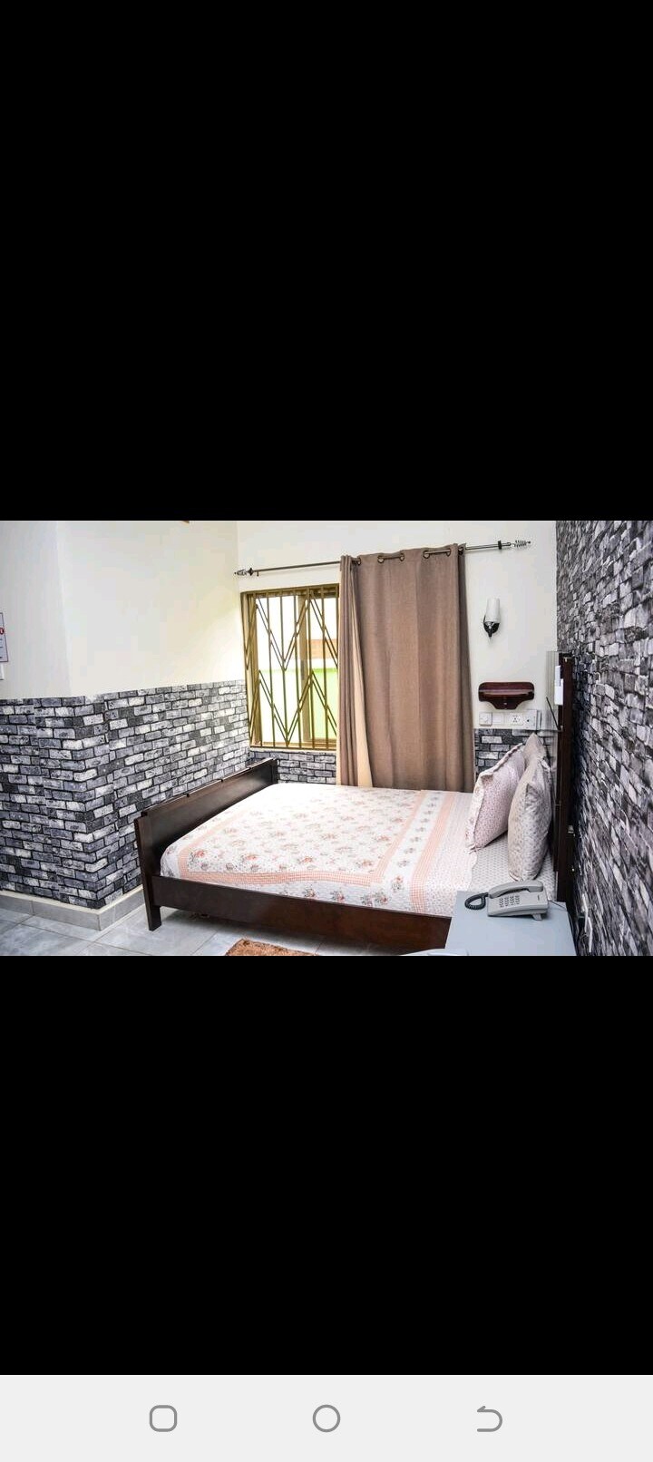 A well-lit bedroom features a spacious bed adorned with a patterned blanket and several pillows. Neutral curtains frame a window, allowing natural light to enter. Textured stone walls create a modern aesthetic, complementing the minimalist design of the room.