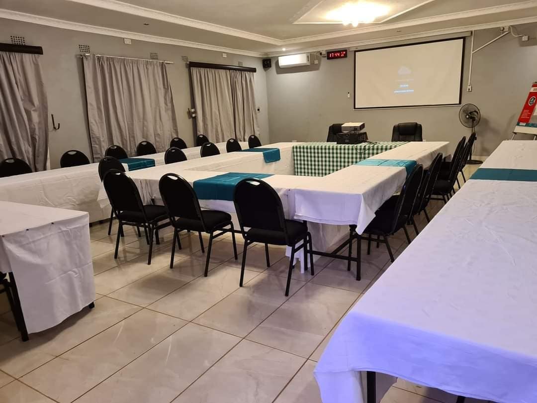 A spacious meeting room is set with multiple tables arranged in a U-shape. Black chairs surround the tables draped in white and teal cloth. Large windows are covered with light gray drapes, and a projector screen is centered at the front of the room.