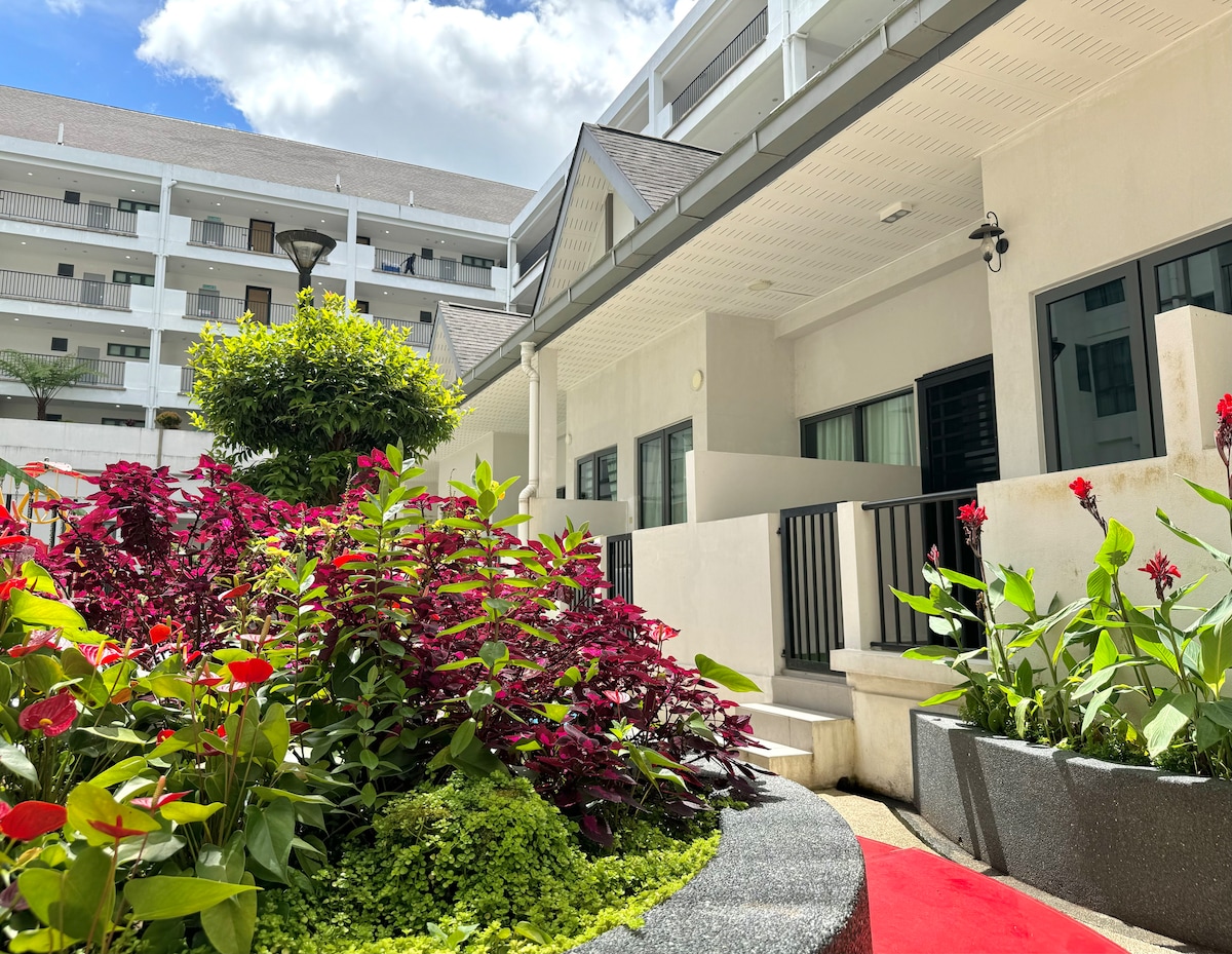 The exterior of the building is framed by vibrant landscaping, featuring lush green plants and colorful flowers. A clear sky and sunlight highlight the welcoming entrance, while the structure's multi-story design is visible in the background.