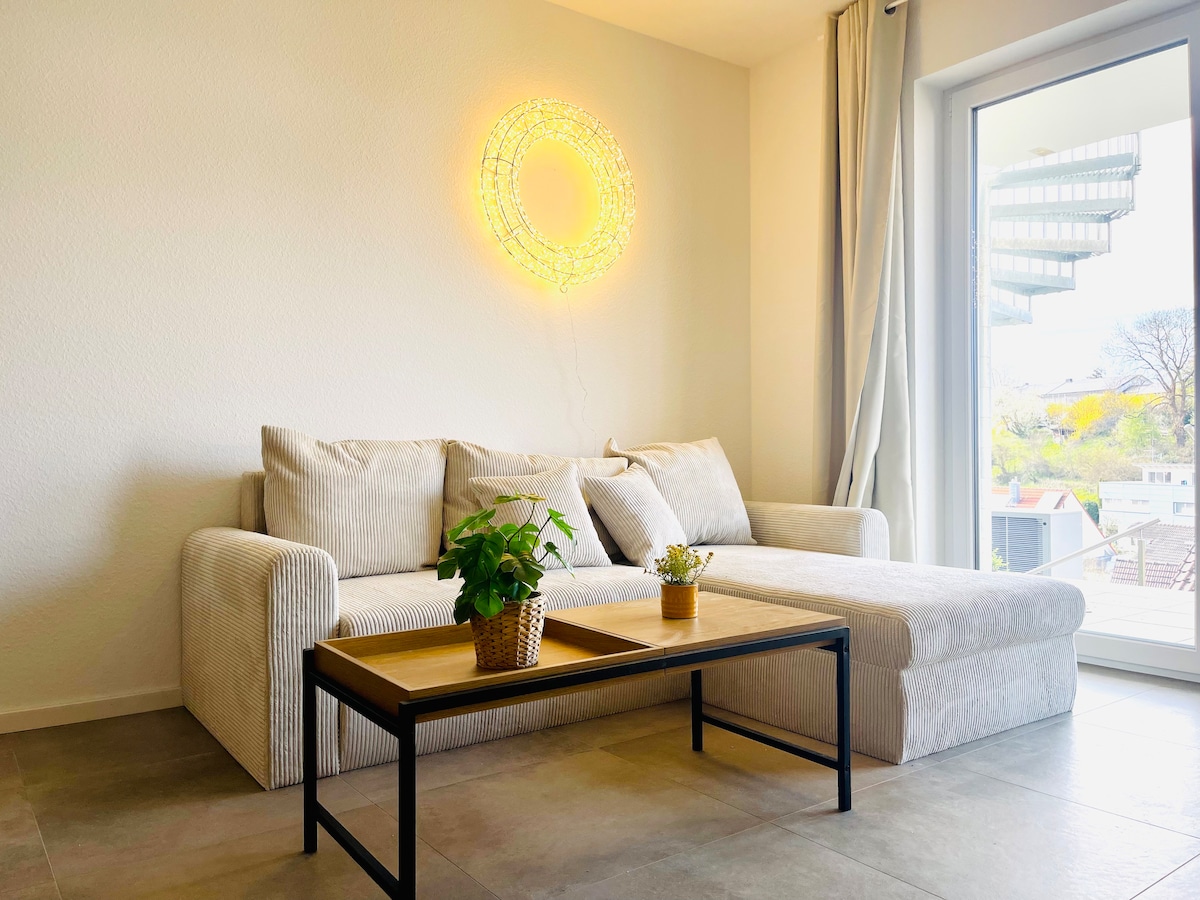 A bright living area features a light-colored couch with a chaise lounge, accompanied by a sleek coffee table. A decorative plant sits in a pot on the table, while a unique wall light casts a soft glow. Large glass doors open to a view outside.