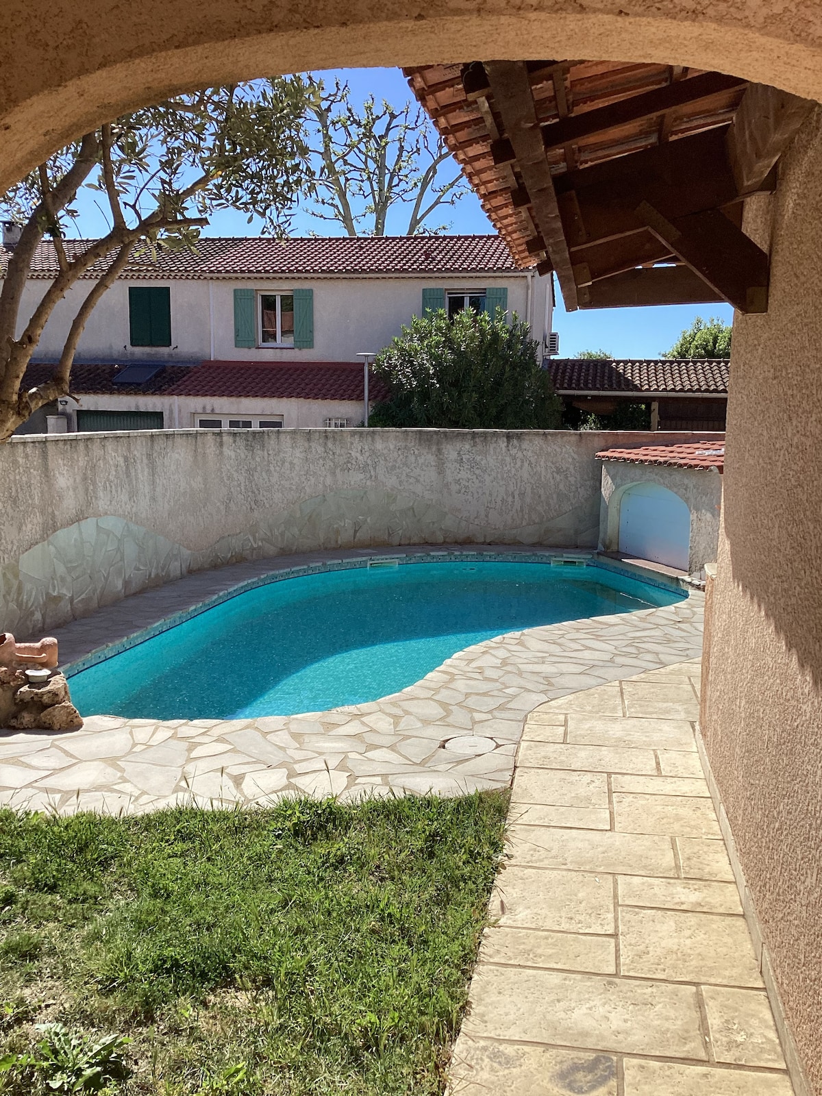 A swimming pool is framed by a stone pathway and grass, positioned under a clear blue sky. The pool features inviting turquoise water and is bordered by a textured wall, with a glimpse of surrounding residential buildings visible in the background.