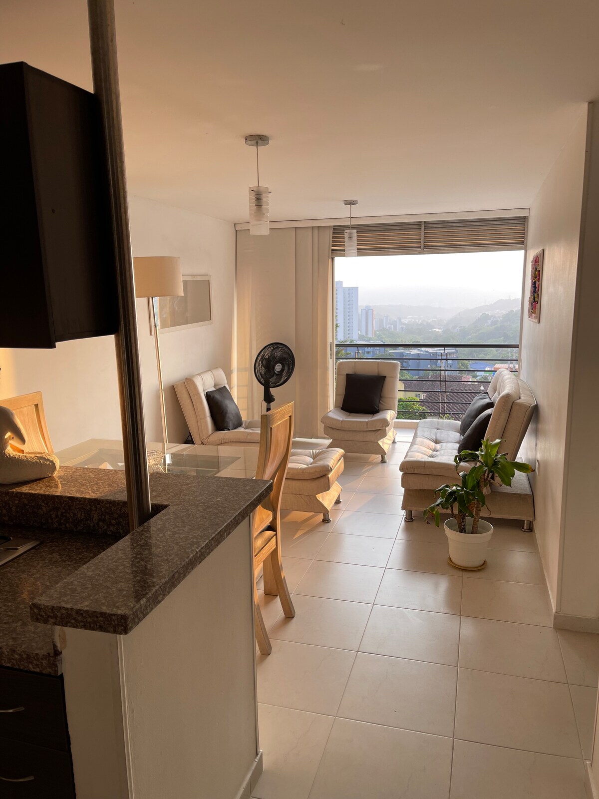 A bright living area features light-colored furnishings, including a two-seat sofa and single chairs positioned around a small table. Natural light floods the space through large windows, revealing a view of the city and surrounding hills. A potted plant adds a touch of greenery.