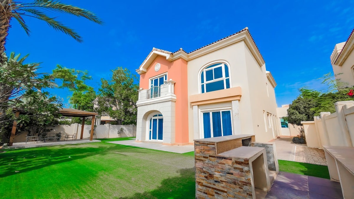 A spacious exterior view of a two-story home is presented, featuring large windows and a soft color palette of light peach and cream. Lush green grass surrounds the property, with palm trees and shrubbery providing natural elements. A stone structure adds texture to the landscape.