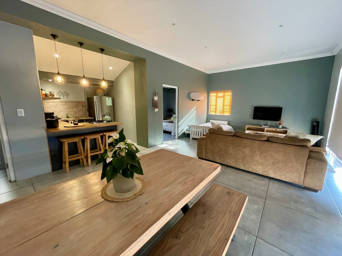 An open plan dining and living area is displayed, showcasing a spacious wooden dining table with bench seating. A comfortable couch faces a wall-mounted television. Soft natural light enters through the windows, highlighting the modern kitchen in the background equipped with stainless steel appliances.