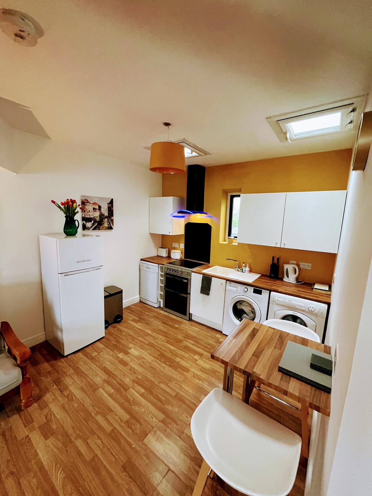 The compact kitchen area is characterized by light wood flooring and yellow walls. It includes a refrigerator, washing machine, and stove, with a small dining table and white chair positioned nearby. Natural light enters through a window above the sink, enhancing the functional space.