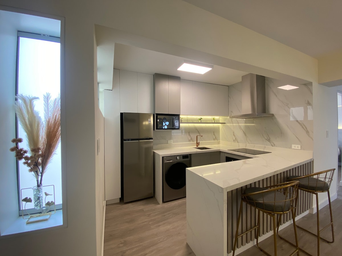 A modern kitchen is showcased with sleek cabinetry and contemporary appliances. A marble countertop extends towards stylish bar seating made of metal and wood. A window displays decorative plants, creating a bright and airy setting.