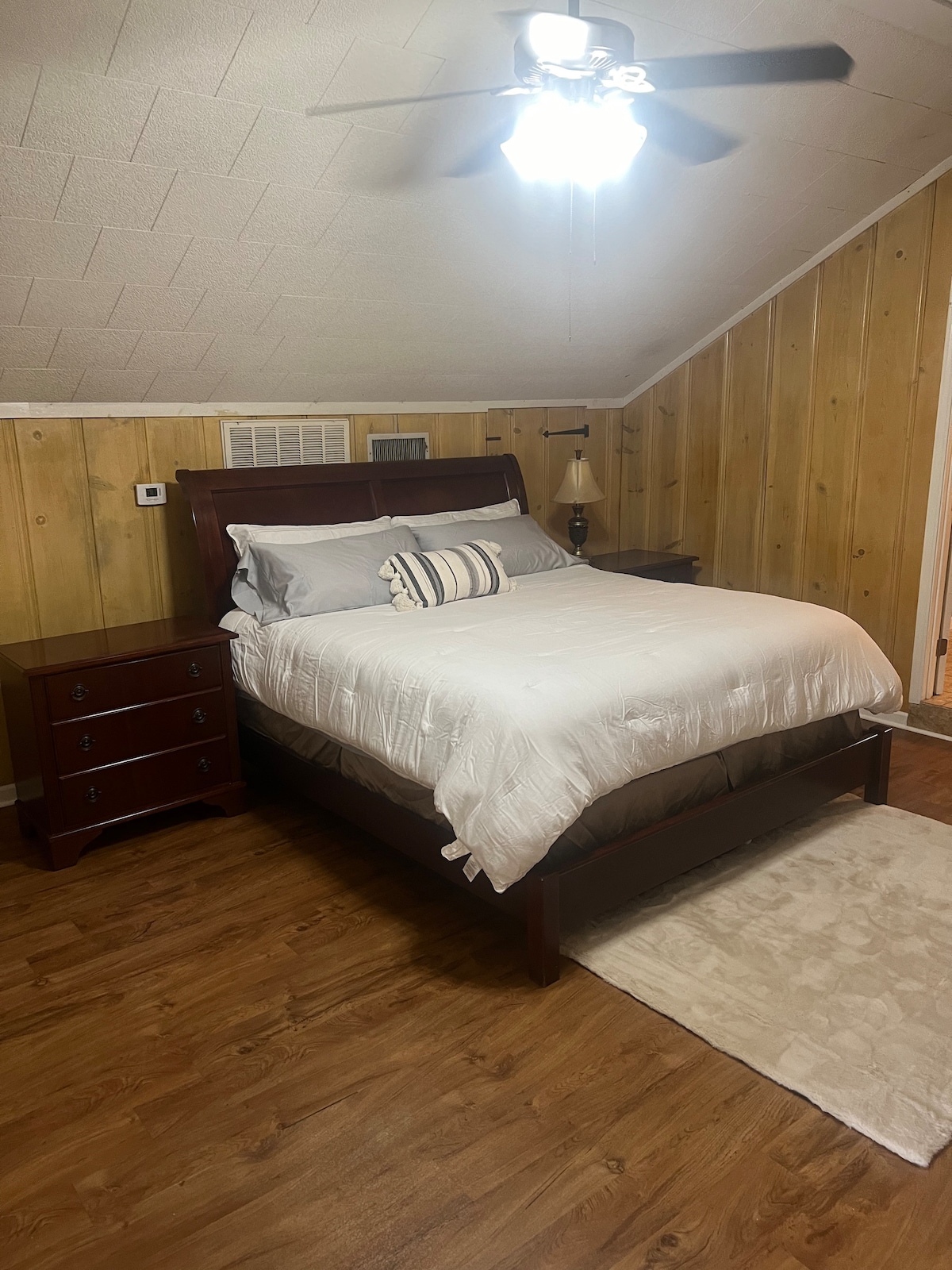 A cozy bedroom is featured with a wooden bed frame and a white comforter. There are two bedside tables, one with a lamp. Wood paneling decorates the walls, and a light rug lies beneath the bed, complementing the warm wooden flooring.