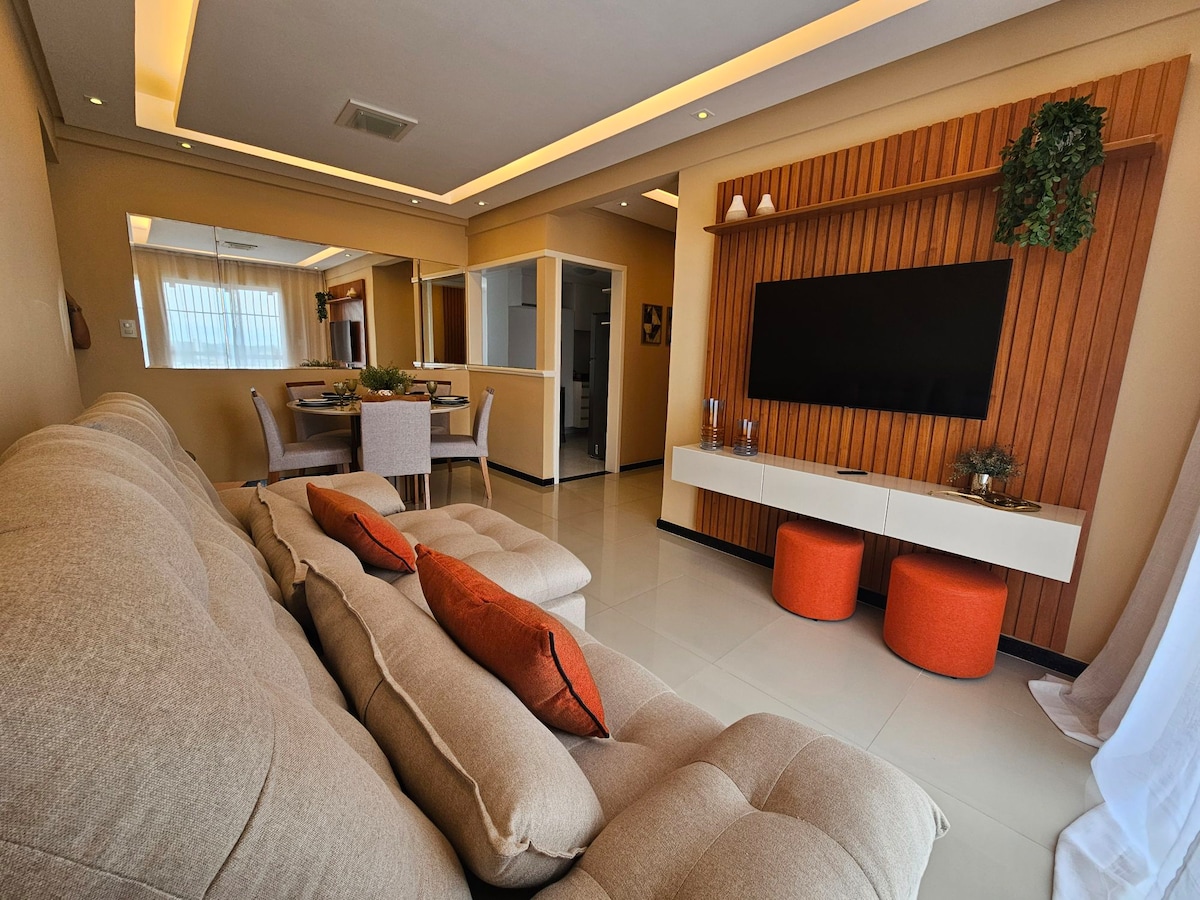 A spacious living area features a comfortable beige sofa with decorative orange cushions facing a wall-mounted television. A light wood panel adds texture to the space. Dining chairs surround a modern table, and the room is illuminated by soft overhead lighting.