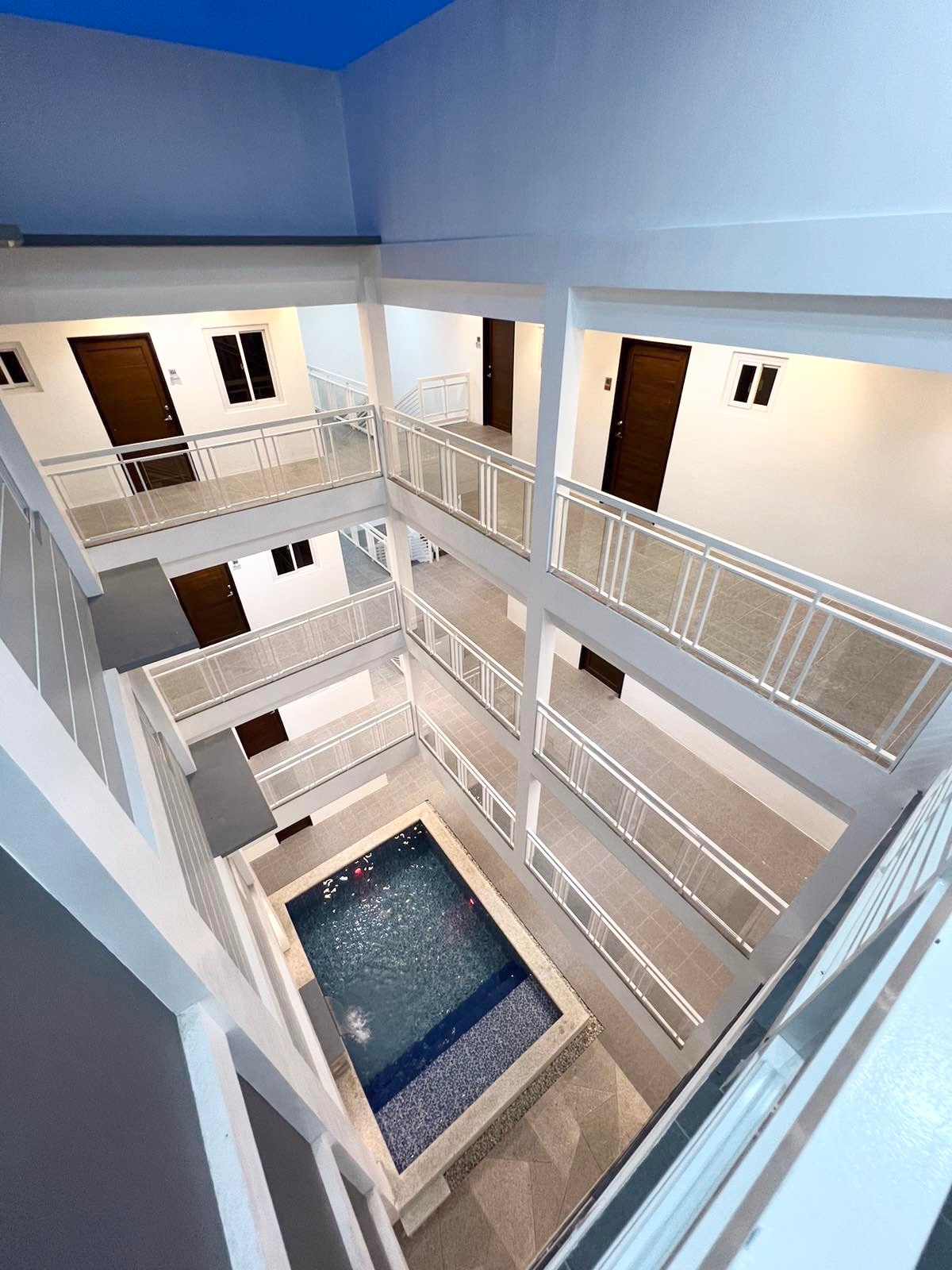An aerial view captures a multi-level building with a central courtyard featuring a pool. The pool is surrounded by a stone deck and is bordered by multiple floors with white railings. Doors are visible leading to individual rooms, enhancing the open atmosphere.