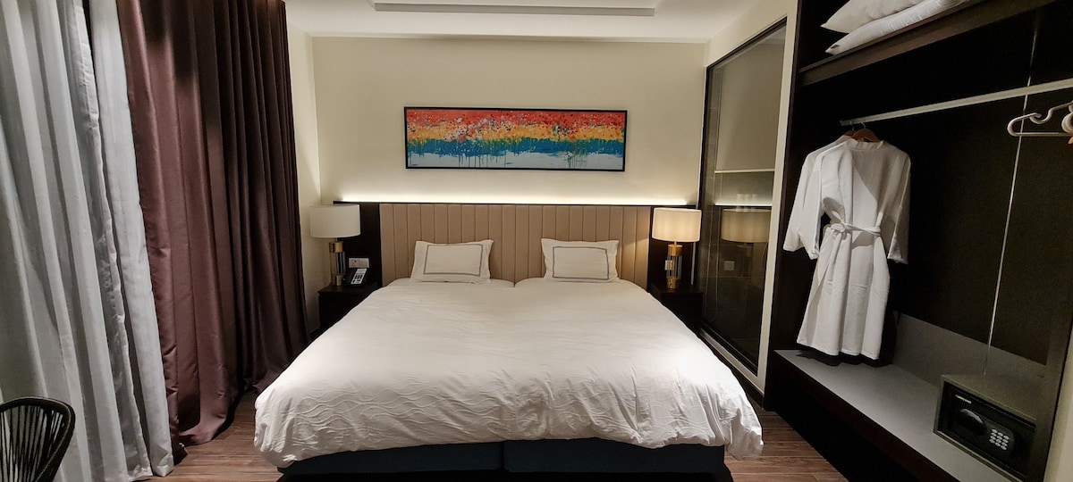 A spacious sleeping area features a large bed dressed in white linens, framed by soft lighting. An abstract artwork hangs above the bed, and nearby storage displays a robe. Soft curtains partially cover a window, complementing the warm color palette.