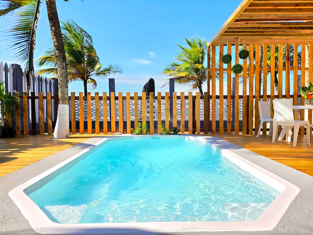 A private pool is surrounded by wooden decking and palm trees, providing a refreshing retreat. Nearby, a shaded area features simple white furniture and hanging plants, creating a serene environment. The clear blue water reflects the sky, while a low wooden fence adds to the secluded feel.