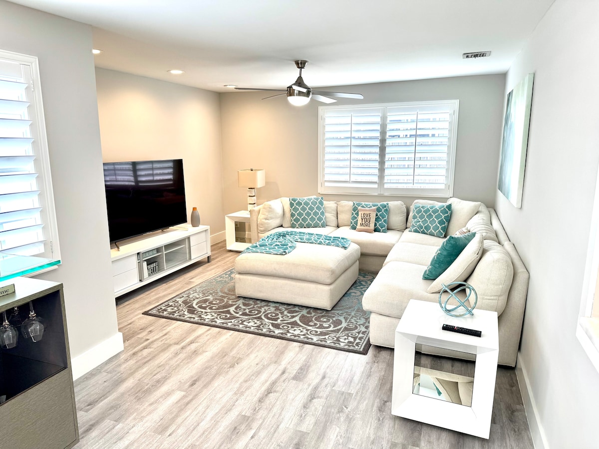 A spacious living area is presented with a large sectional sofa adorned with teal and gray accent pillows. A flat-screen television is mounted on the wall, complemented by a white media console. Natural light enters through the window, situated above a soft area rug on the floor.