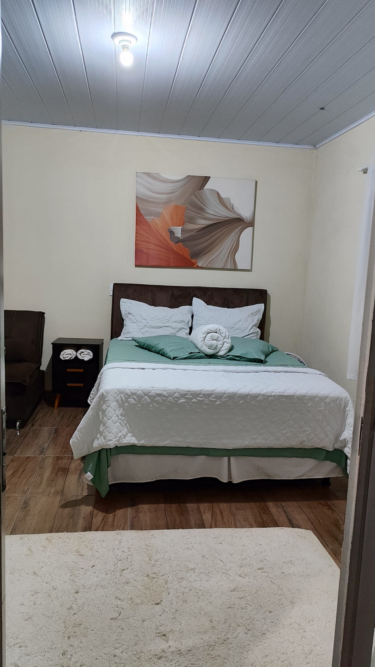 A cozy sleeping area is presented, featuring a spacious bed dressed in soft linens and accented by decorative throw pillows. A piece of wall art adds visual interest, while a small side table holds two clocks. Natural light filters through the nearby window, enhancing the room's warmth.
